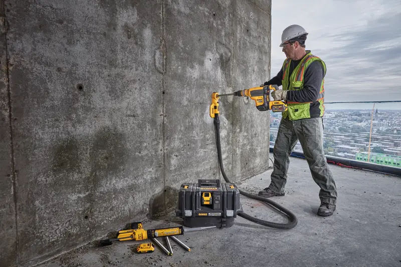 A construction worker wearing a safety vest and helmet is using a DEWALT cordless drill with dust extraction attachment to drill into a concrete wall. The worker is standing on a large building with a cityscape in the background. Several DEWALT tools and a tool case are placed on the ground nearby.